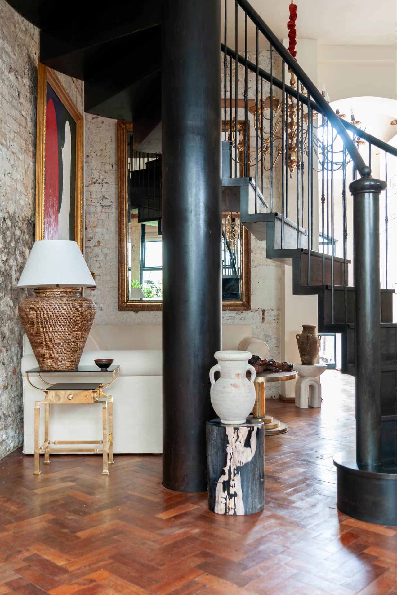 rattan lamp on brass and glass side table by cream sofa and petrified tree stump with terracotta amphora. Steel staircase
