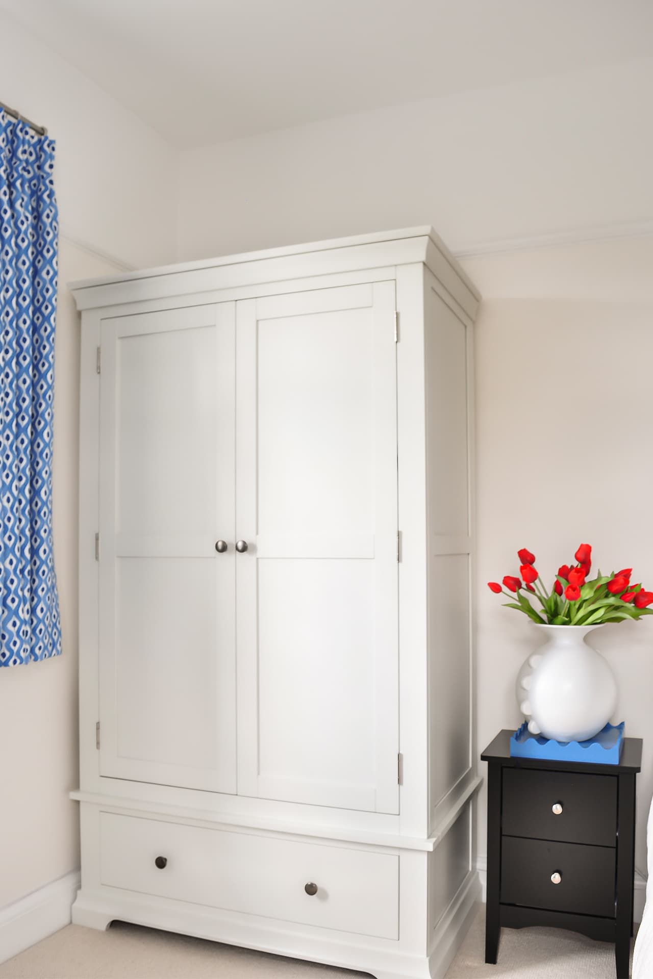 cream bedroom with cream bed, black bedside drawers, blue cushion on bed and blue curtains